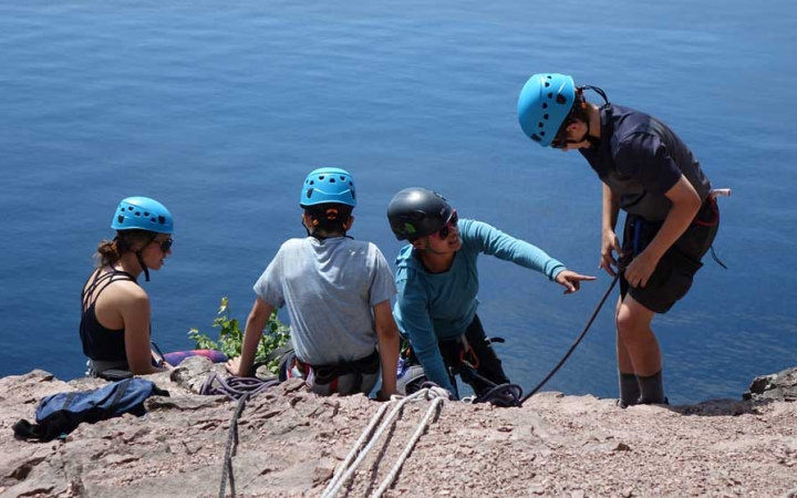 Four people wearing safety gear are secured by ropes near the edge of a cliff over a blue body of water. One person appears to be an instructor, providing guidance to a student.